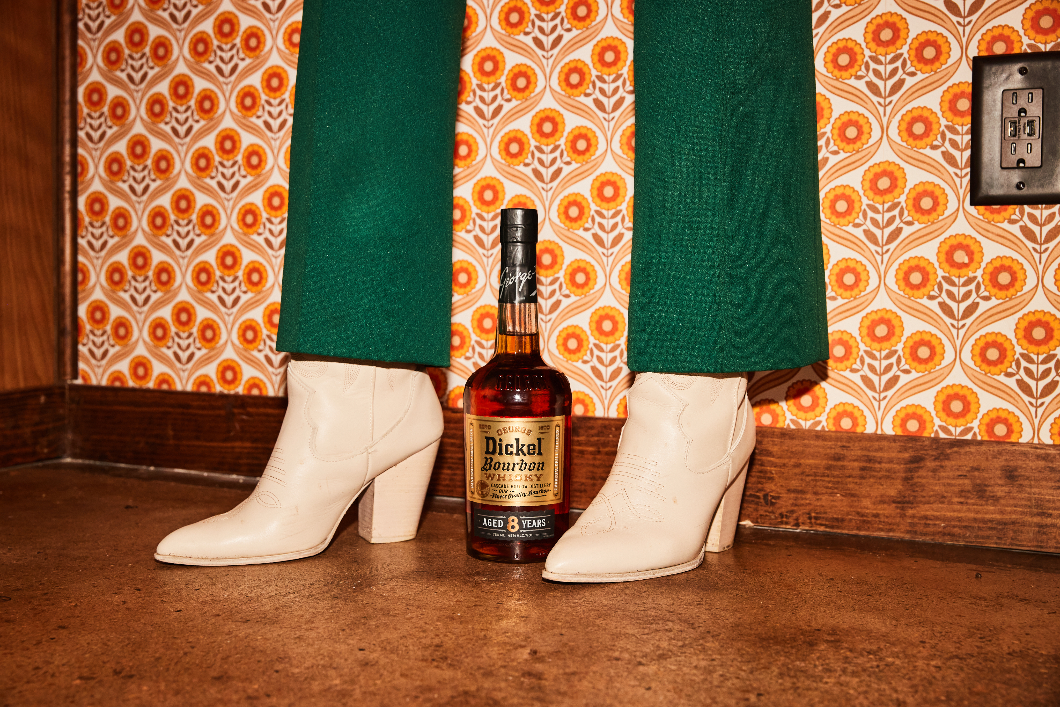 A bottle of Dickel Bourbon sits on the floor between a pair of white heeled boots, next to a wall with orange floral wallpaper and a power outlet.