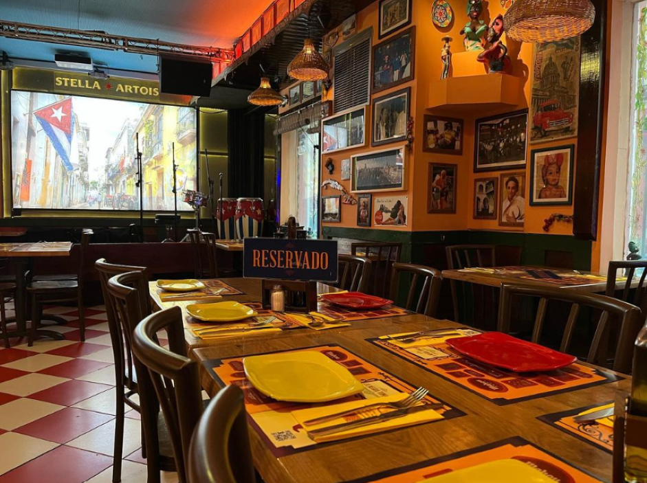 La foto muestra el interior de La Negra Tomasa. Hay mesas de madera preparadas para comer, con platos amarillos y servilletas rojas. El suelo tiene un patrón de cuadros rojos y blancos. En las paredes, hay varias decoraciones, incluyendo banderas.