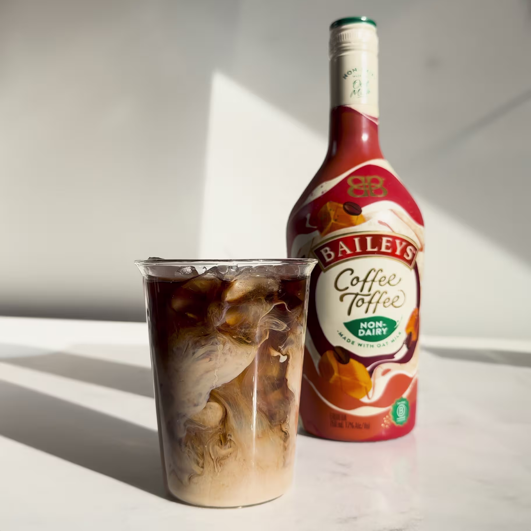 A glass of iced coffee with cream swirling inside is placed in front of a bottle of Baileys Coffee Toffee Non-Dairy creamer on a white surface.