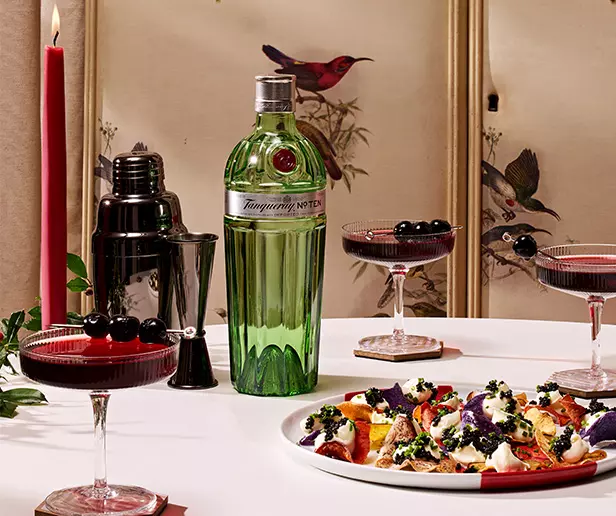 A bottle of Tanqueray gin, a cocktail shaker, three cocktails with cherries, a red candle, and a plate of food are arranged on a table with a bird-themed screen in the background.