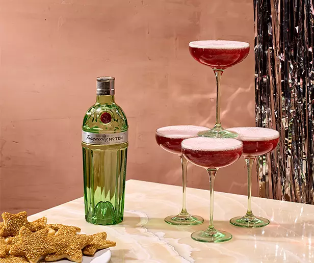 A bottle of Tanqueray No. Ten gin next to a pyramid of four coupe glasses filled with a red cocktail, with star-shaped cookies on a plate in the foreground.