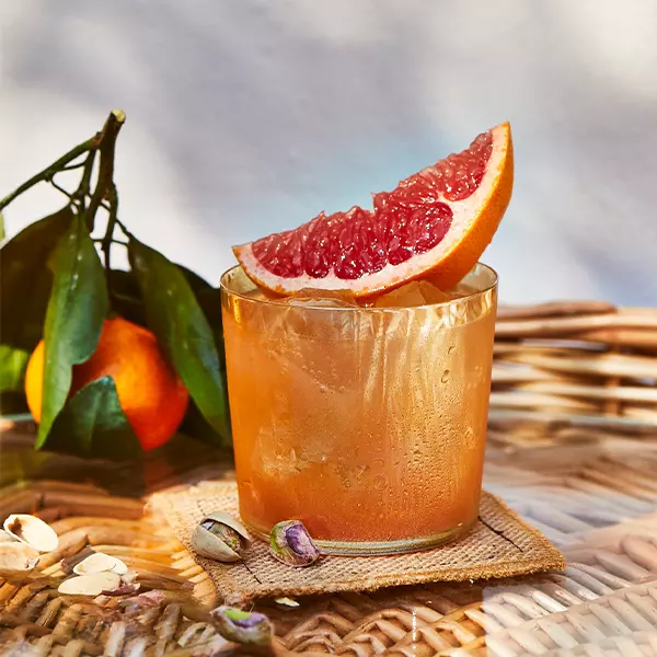 A glass of iced grapefruit drink garnished with a grapefruit slice sits on a wicker table next to a tangerine with leaves and a few scattered pistachios.