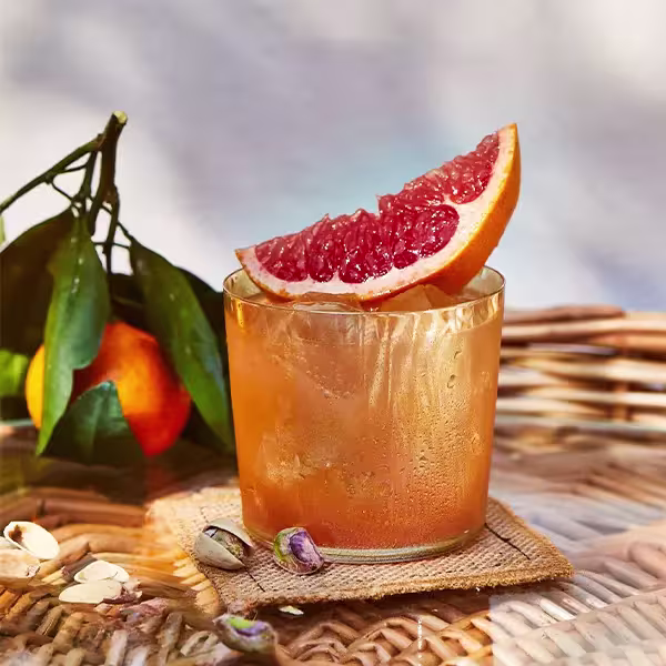 A glass of iced grapefruit drink garnished with a grapefruit slice sits on a wicker table next to a tangerine with leaves and a few scattered pistachios.