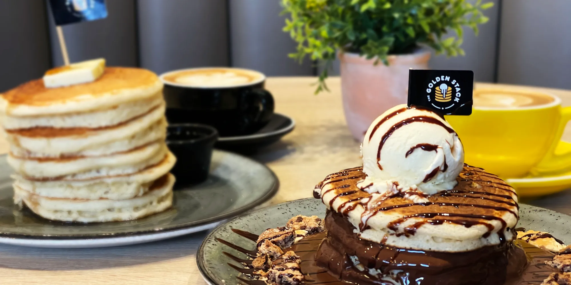 Platos de pancakes en The Golden Stack, uno con sirope y mantequilla, otro con helado y chocolate, acompañados de cafés y fondo con planta decorativa.