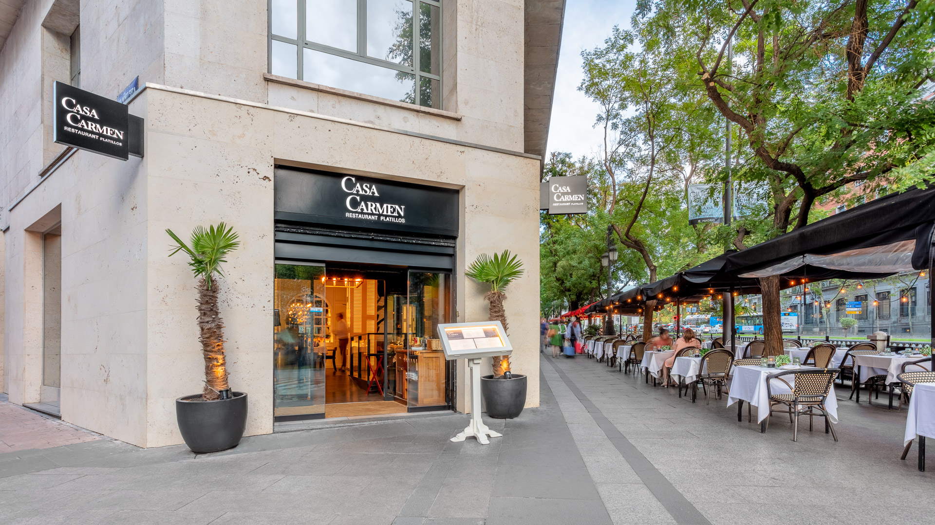 Fachada de Casa Carmen Madrid, con su entrada iluminada y terraza exterior de mesas con manteles blancos, ideal para disfrutar al aire libre en un ambiente acogedor