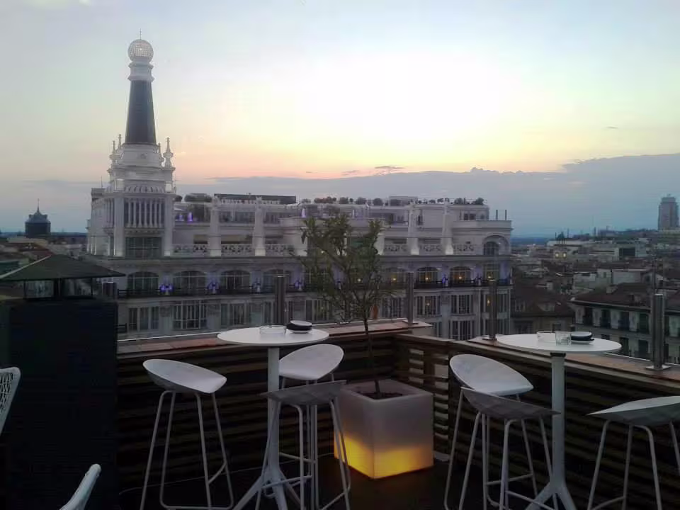 Vista desde la terraza de Club Argo al atardecer con mesas altas, sillas modernas y un paisaje urbano destacado por un edificio emblemático iluminado.