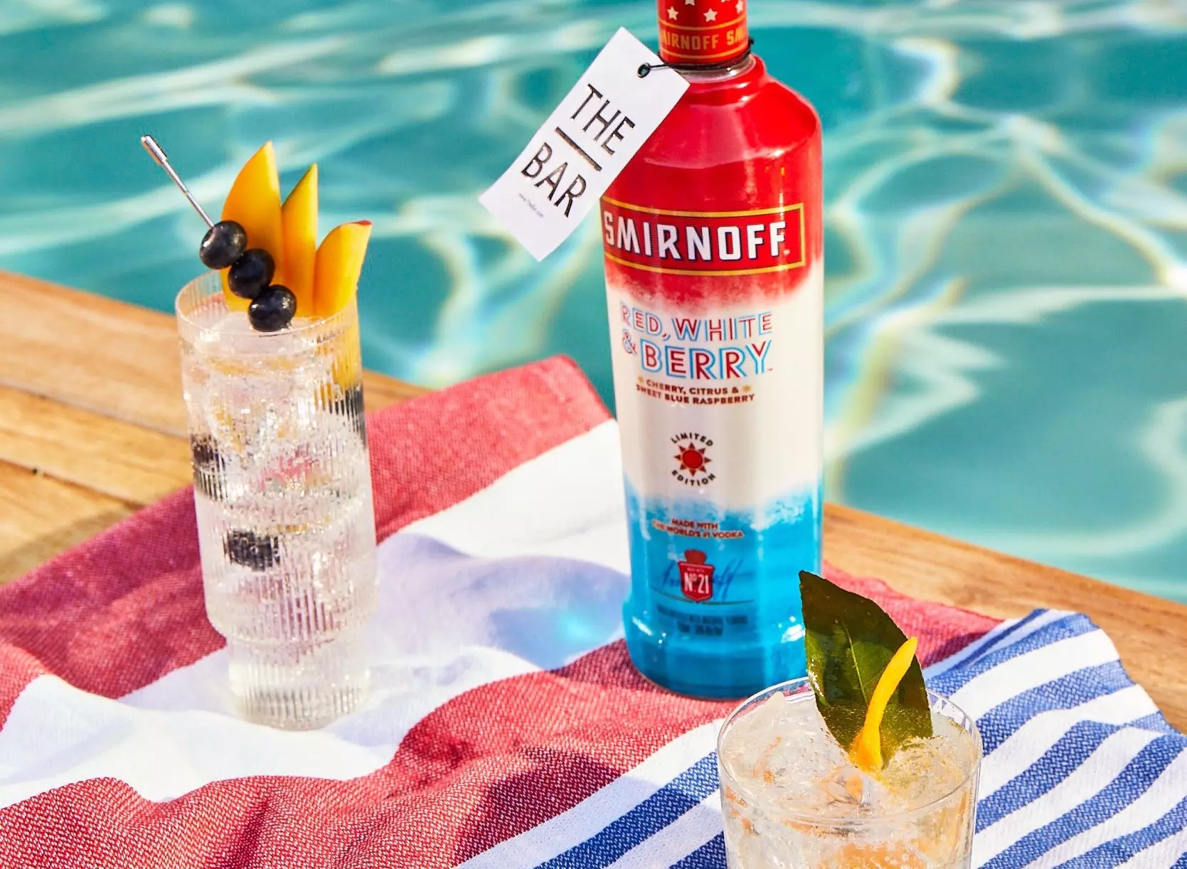 A bottle of Smirnoff Red, White & Berry vodka stands on a striped towel by a pool, next to two cocktails garnished with fruit and a tag reading THE BAR.