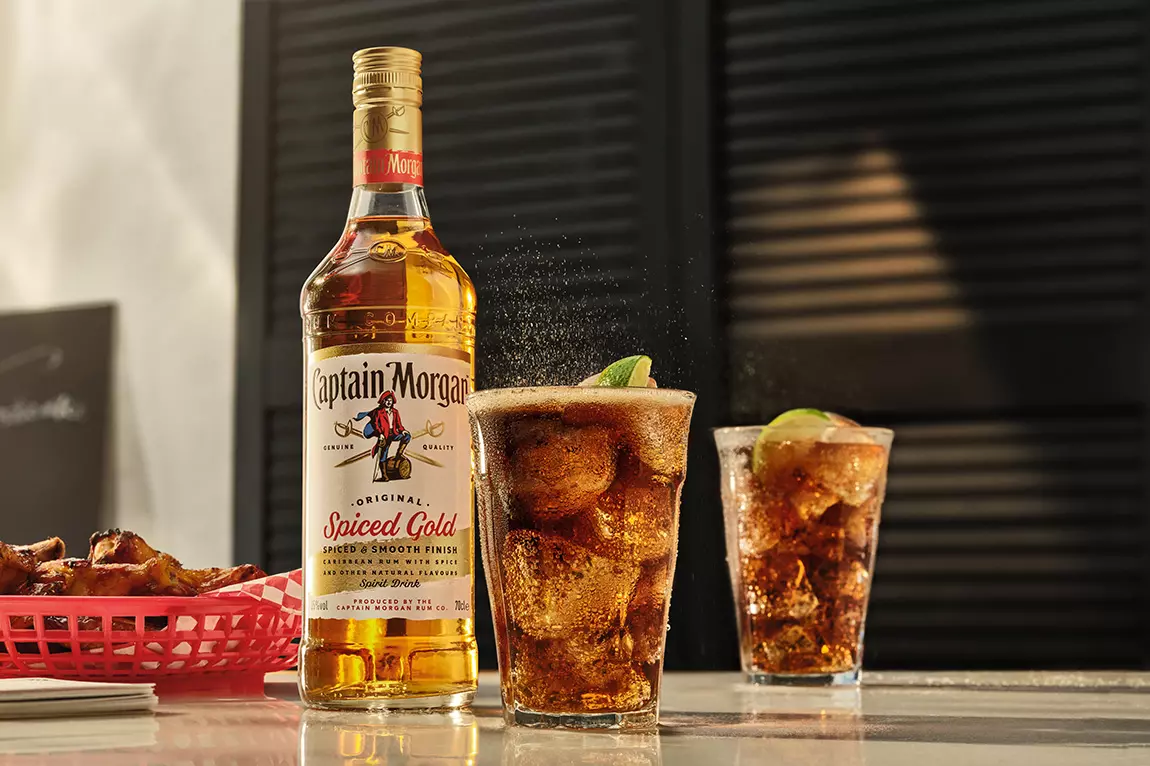 A bottle of Captain Morgan Spiced Gold rum stands next to two glasses of rum and cola with ice and lime wedges on a counter. A basket of food is partially visible in the background.