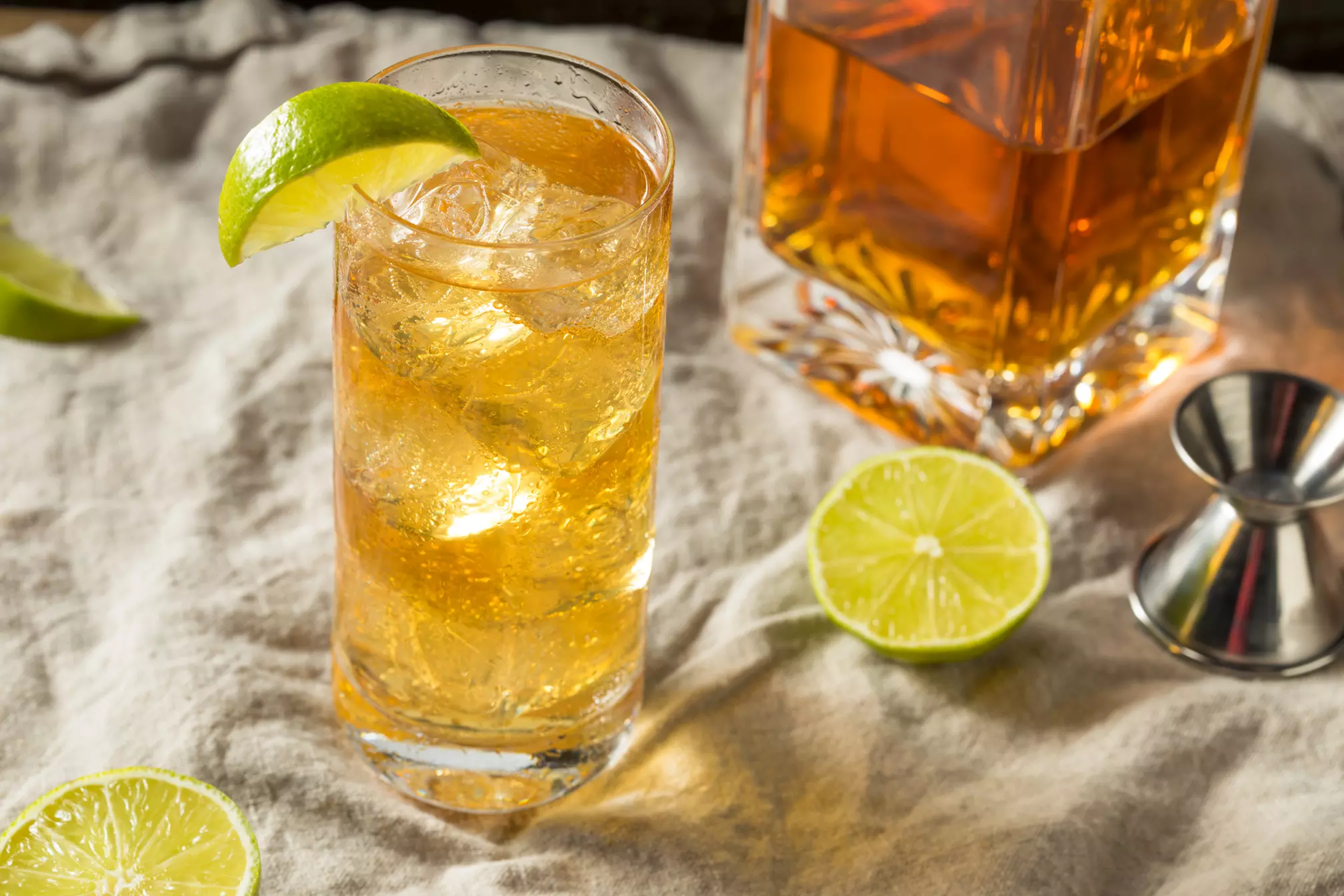 A highball glass filled with a brown cocktail, ice, and a lime wedge, sits on a cloth next to lime slices, a metal jigger, and a decanter of amber liquid.