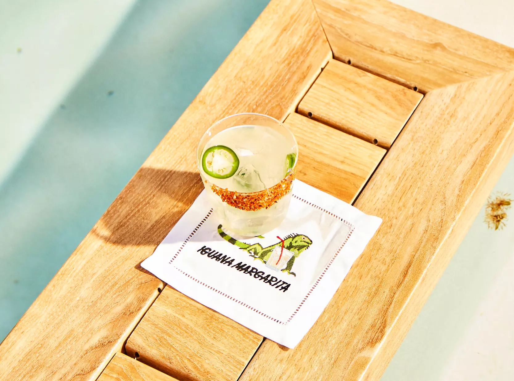 A margarita garnished with lime and jalapeño sits on a napkin labeled IGUANA MARGARITA atop a wooden table by a pool.