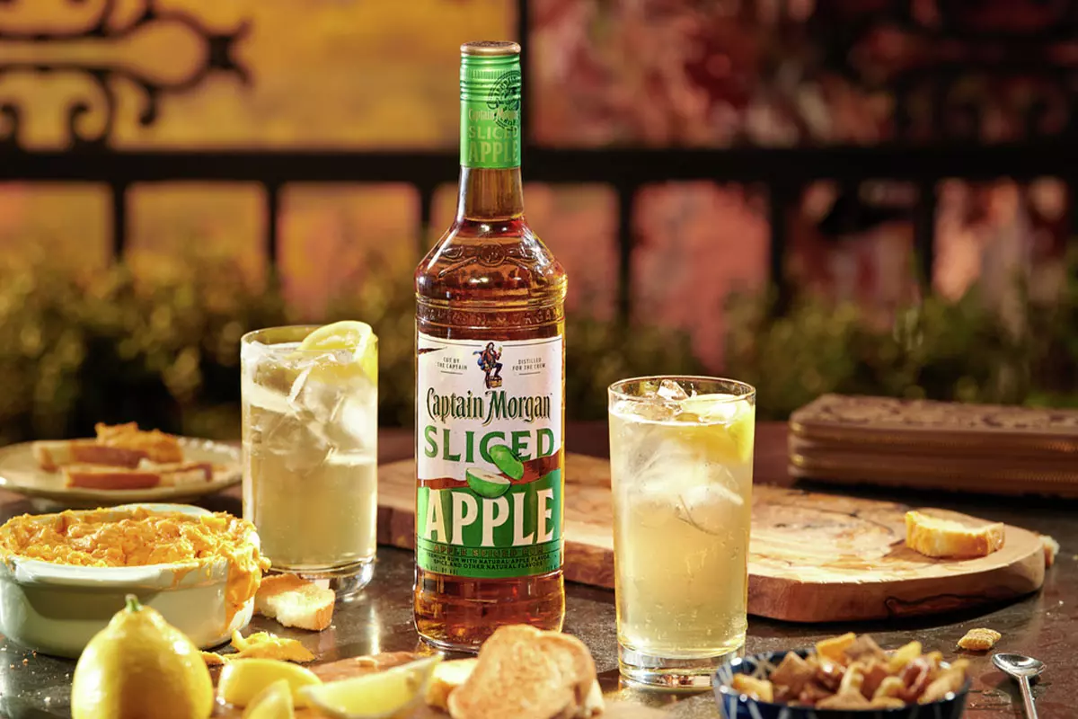 A bottle of Captain Morgan Sliced Apple rum is placed on a table with two cocktails, lemon slices, bread, and a bowl of dip.