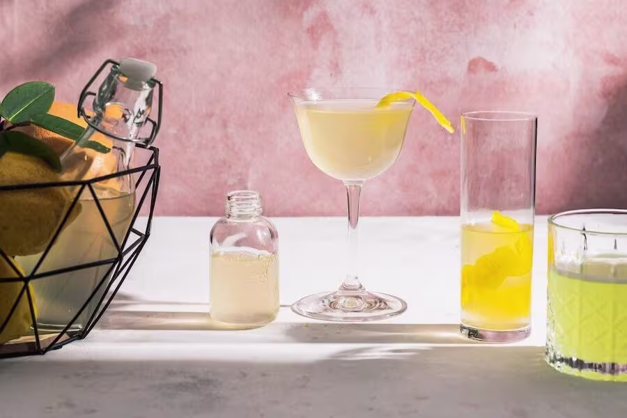 A selection of citrus drinks in various glasses sits on a white surface, with a wire basket of lemons and a small bottle of juice nearby, against a pink backdrop.