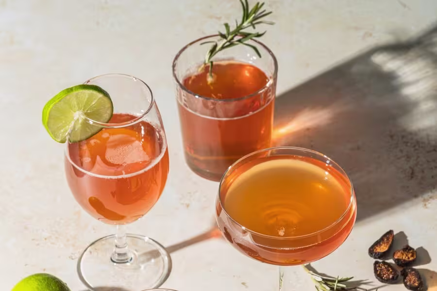 Three cocktails in different glasses, one garnished with a lime slice, another with a rosemary sprig, are set on a light surface with small dried fruit pieces nearby.