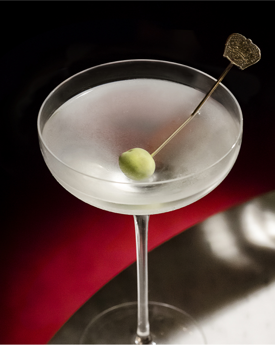 A clear martini cocktail in a coupe glass, garnished with a single green olive on a metal pick, set against a dark background.