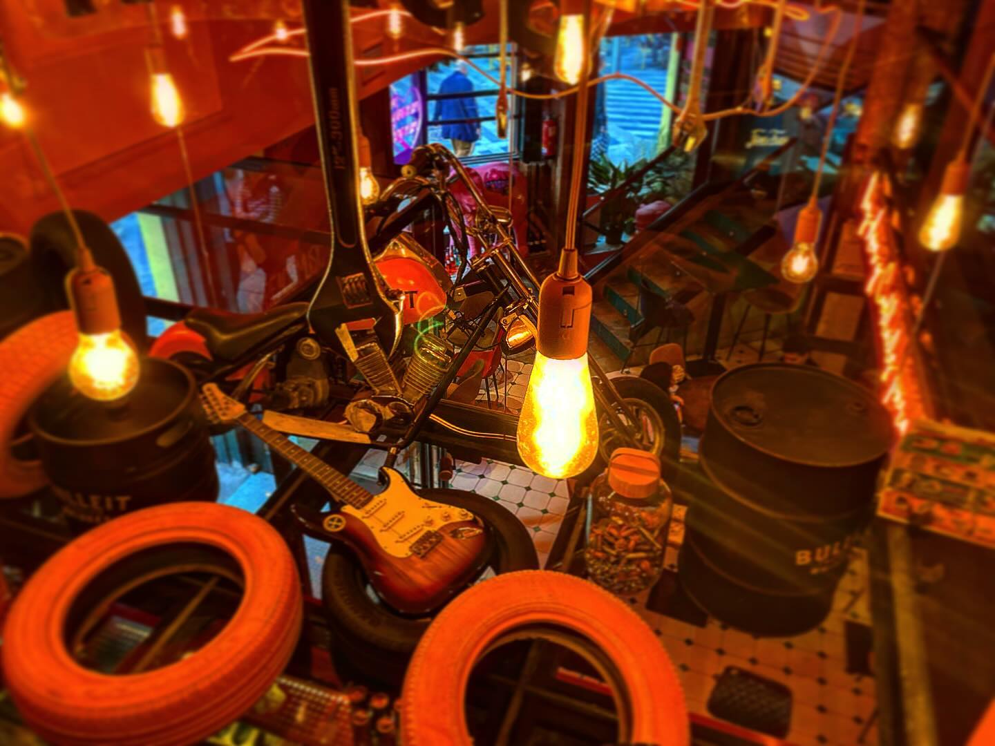 La foto muestra un espacio interior ecléctico y vibrante,del Love Cocktail. Los elementos más llamativos son las numerosas bombillas colgantes de diferentes formas y tamaños, que emiten un brillo cálido. Debajo de las luces, hay una variedad de objetos.