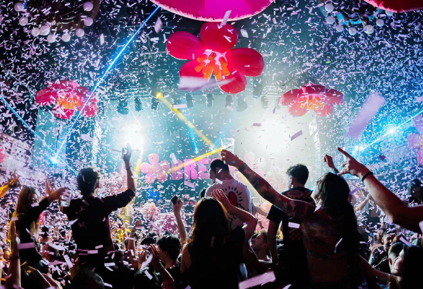 Fiesta en Sala Apolo, con confeti flotando en el aire, decoraciones florales y un público vibrante disfrutando del espectáculo bajo luces brillantes"