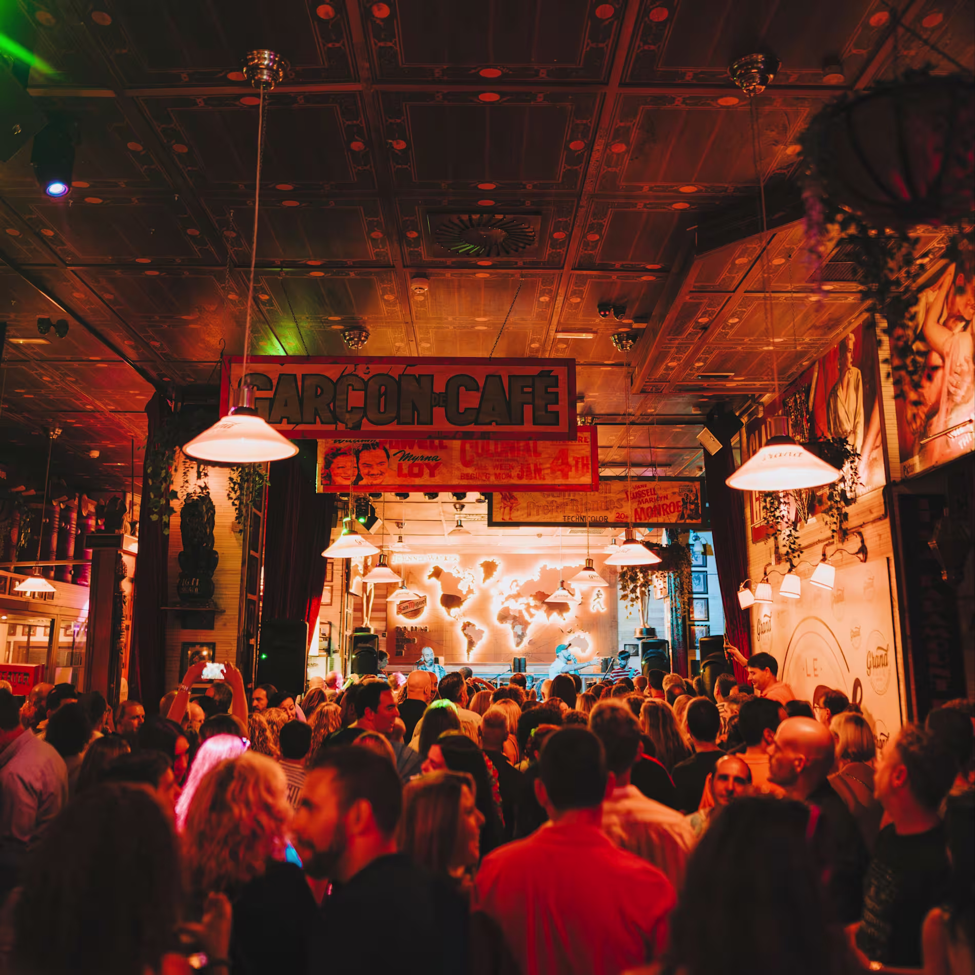 La imagen muestra el interior de un bar o café lleno de gente. El lugar tiene una iluminación cálida y ambiental, decorado con varios elementos colgantes.