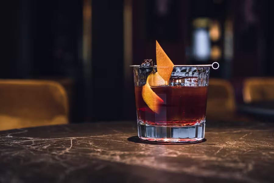 A glass of dark cocktail garnished with an orange peel and a cherry on a cocktail pick, placed on a marble table.