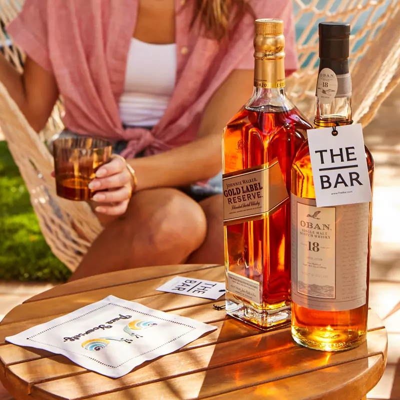 Two bottles of whisky and a glass sit on a wooden table outdoors; a person seated in a hammock holds a drink in hand.