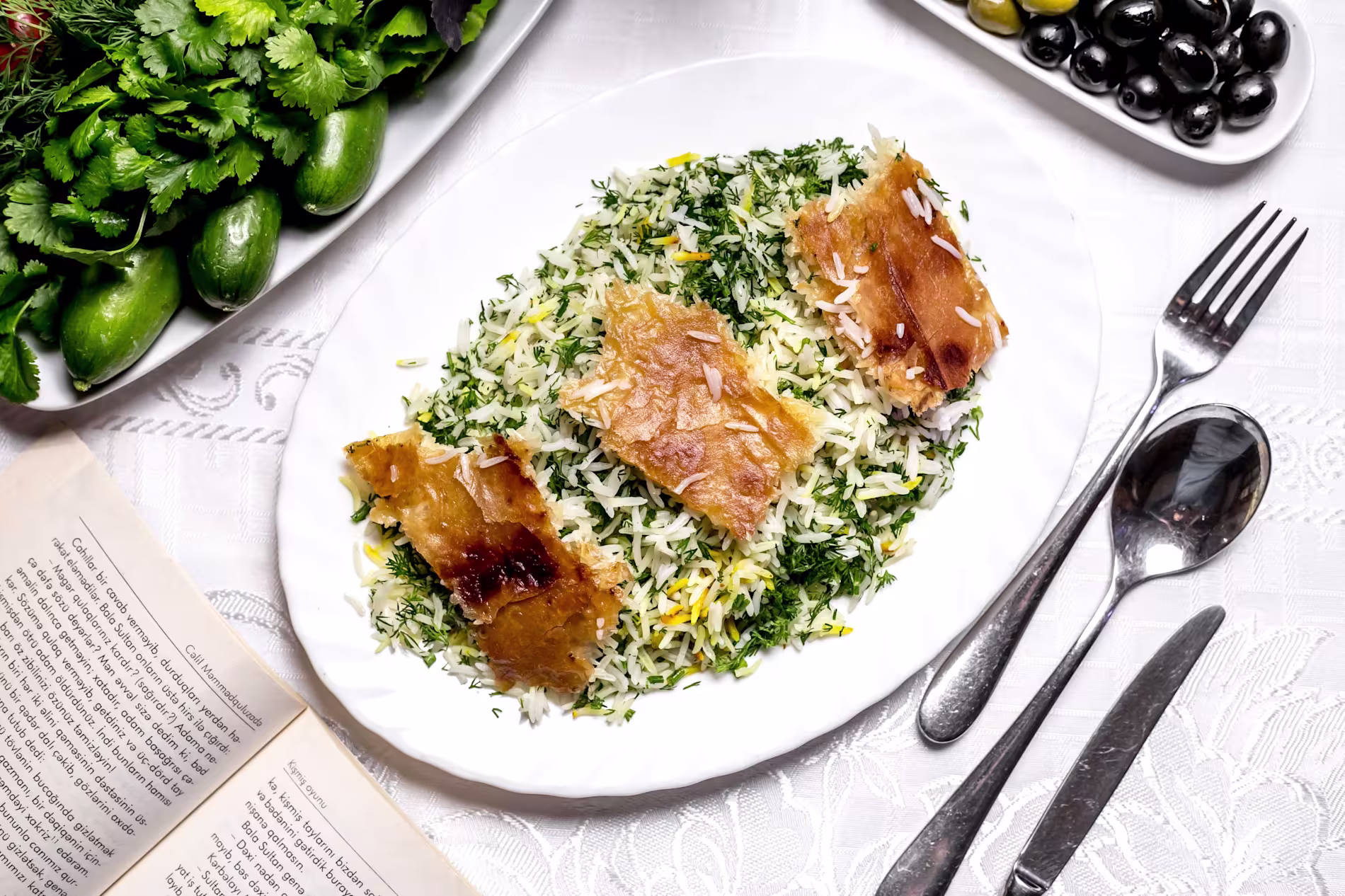 A plate of herbed rice with three pieces of crispy tahdig, served on a white tablecloth with a side of fresh herbs, cucumbers, black olives, and an open book nearby.