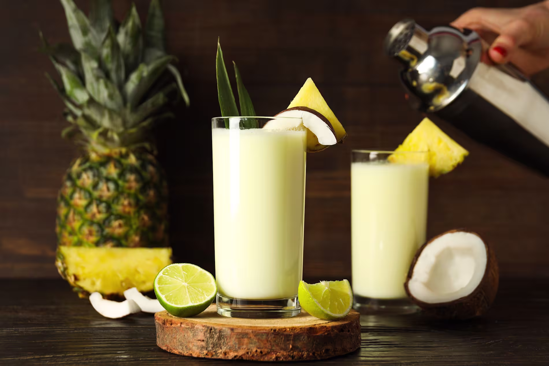 Two glasses of piña colada with pineapple and coconut garnish, surrounded by fresh pineapple, lime, and coconut pieces; a hand pours from a cocktail shaker.