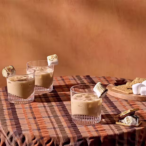 Three glasses of iced coffee are garnished with toasted marshmallows, set on a plaid tablecloth with graham crackers, marshmallows, and chocolate nearby.
