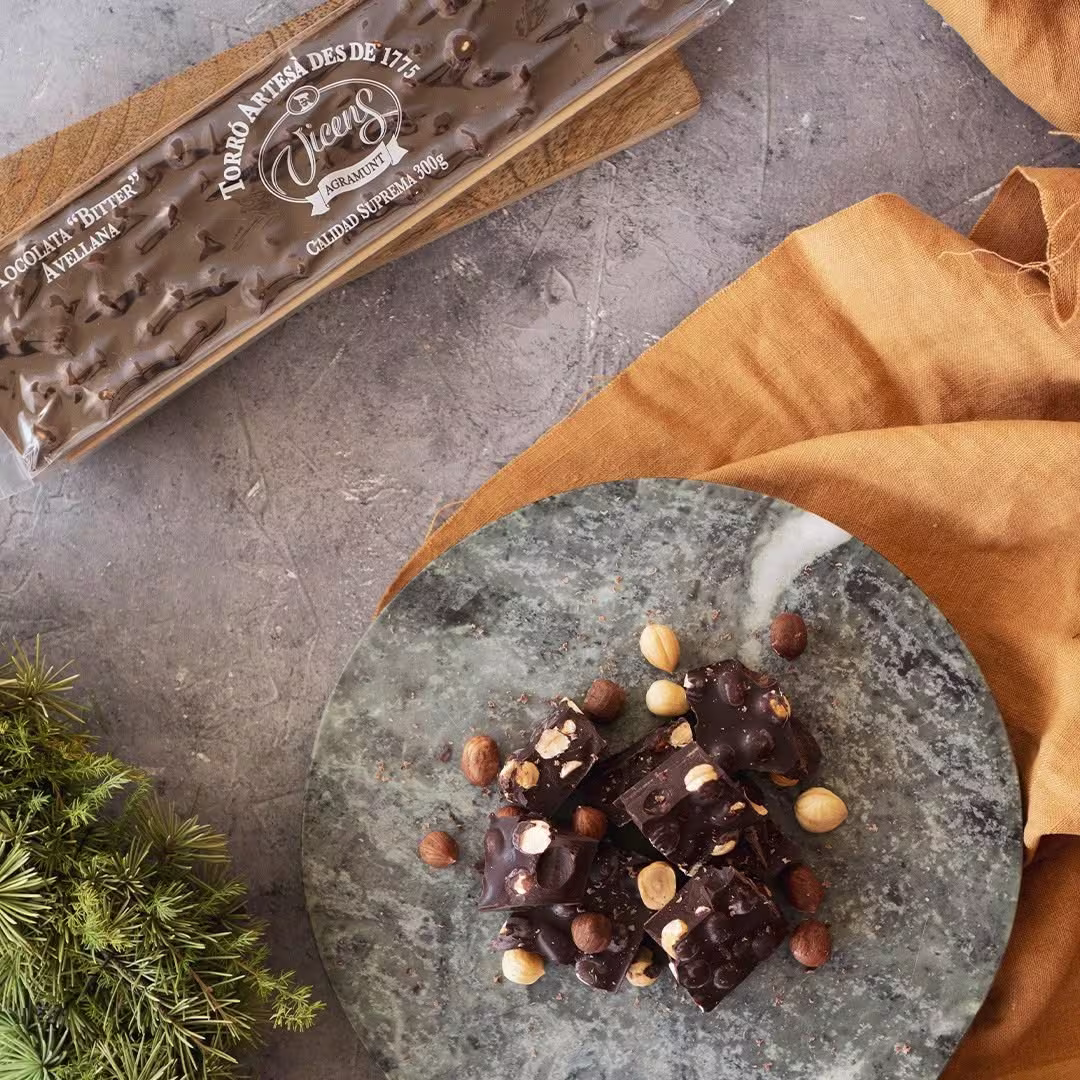 Trozos de turrón de chocolate bitter con avellanas sobre un plato de mármol verde, junto a su tableta original de Torrons Vicens envuelta y una base decorativa de tela mostaza.