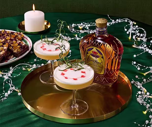 A bottle of Crown Royal whisky and two cocktails with rosemary garnish sit on a gold tray, next to a candle and a plate of brownies on a green tablecloth.