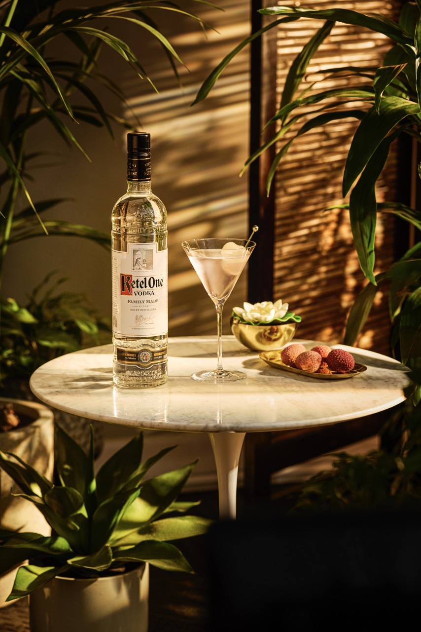 A bottle of Ketel One vodka, a martini glass with a garnish, and a plate of lychees sit on a round marble table surrounded by green plants.