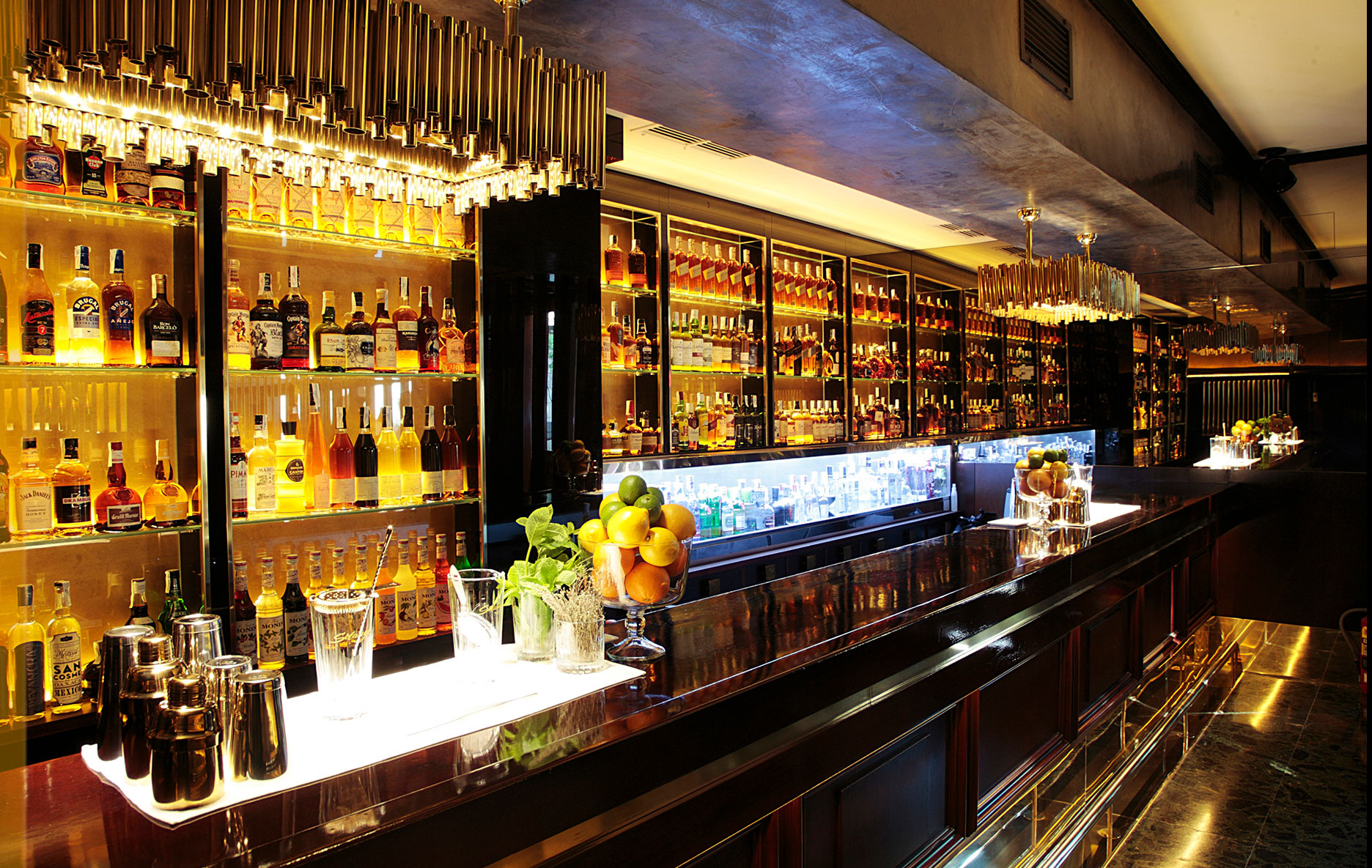 Solange Cocktails presenta una barra sofisticada con estantes iluminados repletos de licores premium y detalles dorados que resaltan su elegancia.