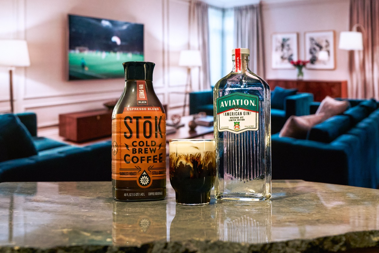 A bottle of Stok cold brew coffee, a glass with a dark beverage, and a bottle of Aviation American Gin on a marble counter with a living room and TV in the background.