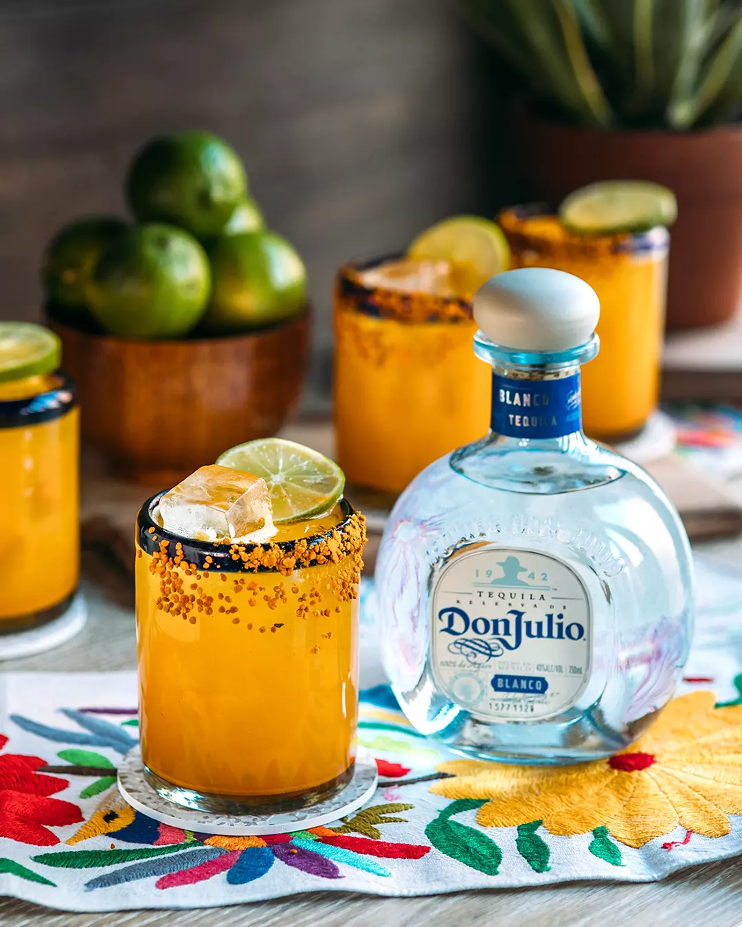 A glass of orange cocktail with ice and a lime slice sits beside a bottle of Don Julio tequila, with limes and similar drinks in the background on a colorful embroidered cloth.