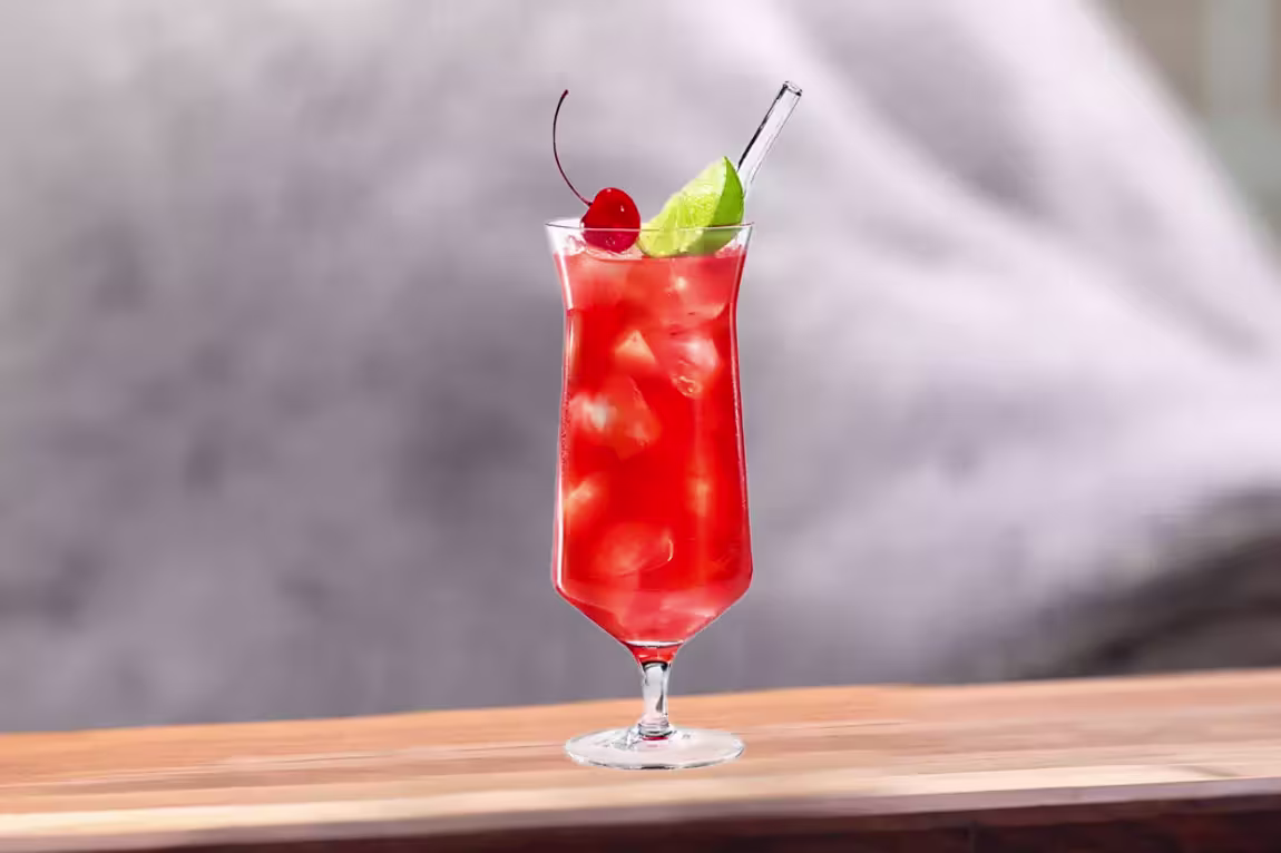 A tall glass filled with red iced beverage, garnished with a cherry, a lime wedge, and a clear straw, placed on a wooden surface.
