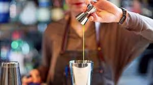 Person pours liquid from a jigger into a cocktail shaker, with bar tools and bottles blurred in the background.