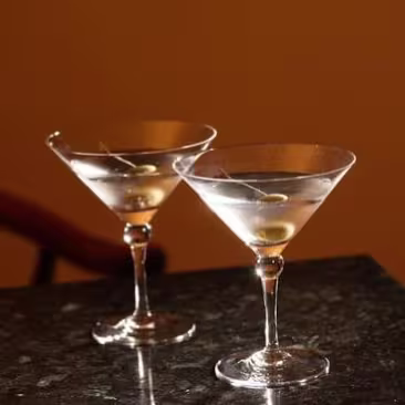 Two martini glasses with olives on picks sit on a bright red surface, casting shadows.