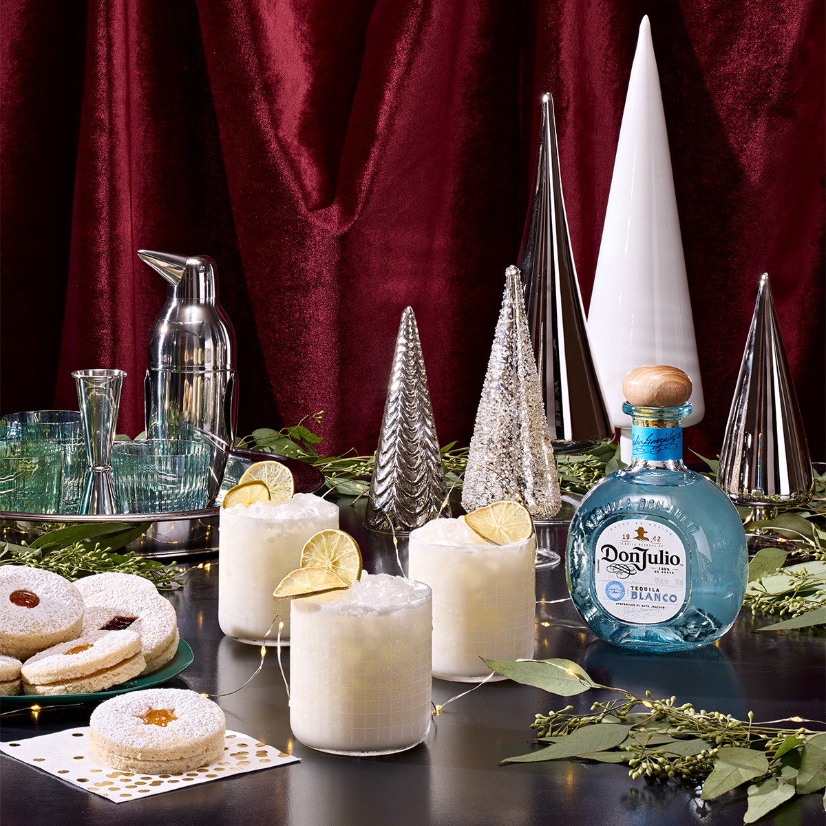 A festive table setting with three creamy cocktails garnished with lime, a bottle of Don Julio tequila, assorted cookies, glasses, a shaker, and silver decorative trees against a red curtain.