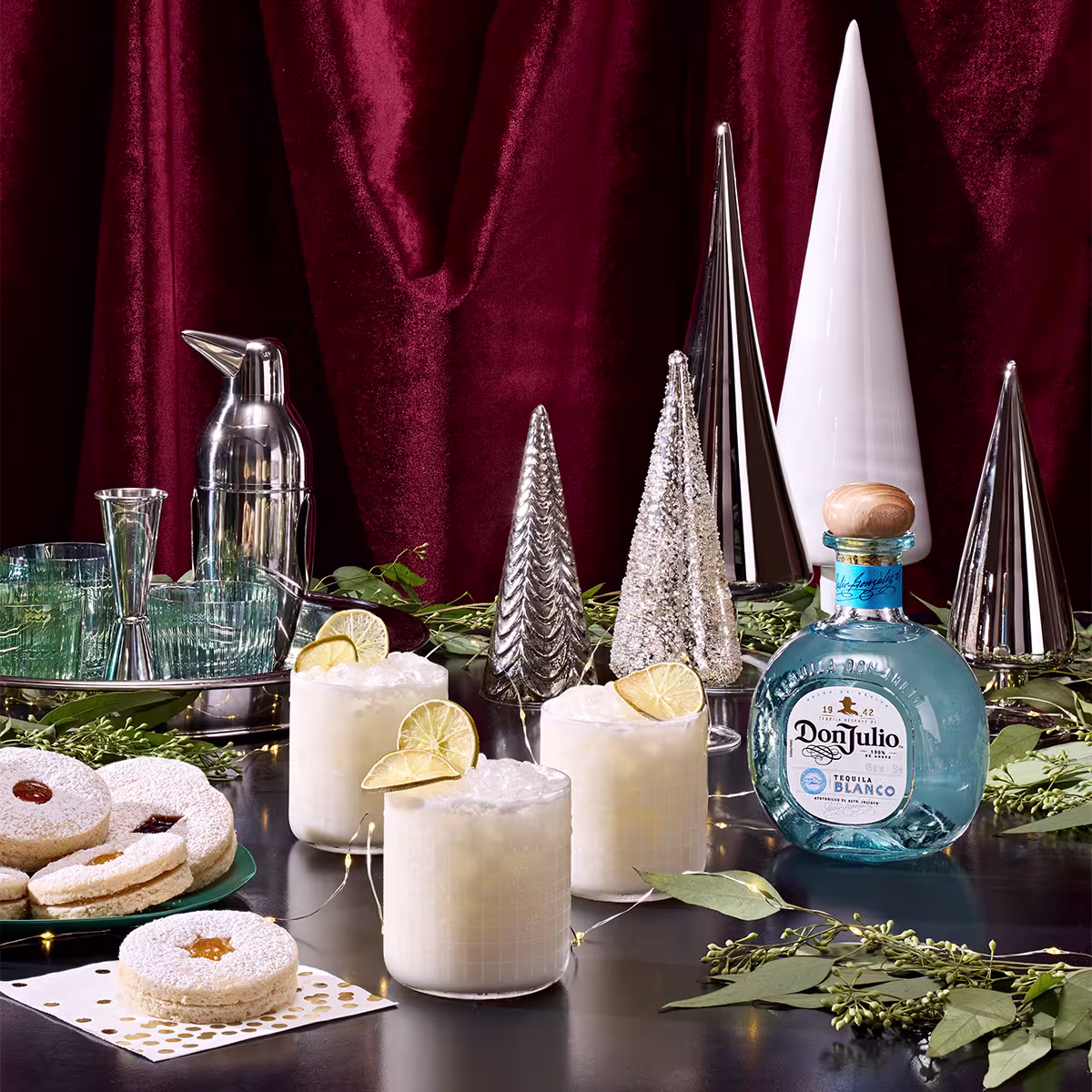 A festive table setting with three creamy cocktails garnished with lime, a bottle of Don Julio tequila, assorted cookies, glasses, a shaker, and silver decorative trees against a red curtain.