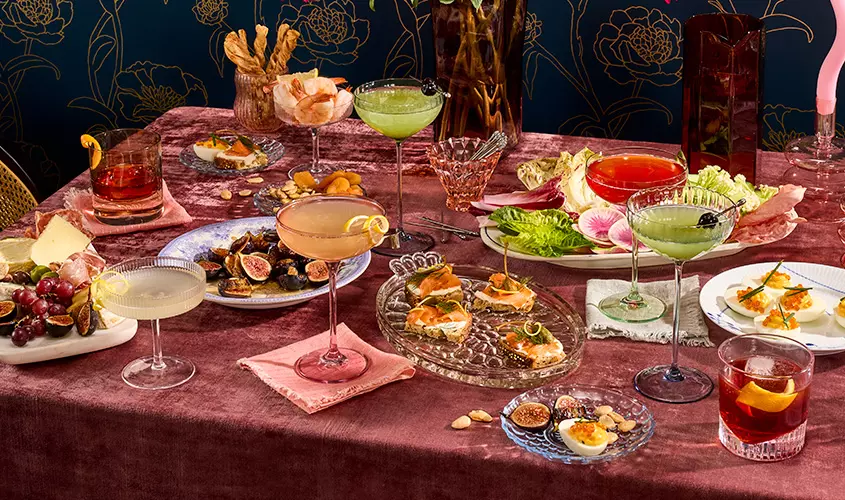 A table set with assorted cocktails, hors doeuvres, cheese, grapes, figs, and leafy greens on elegant dishes and glassware.