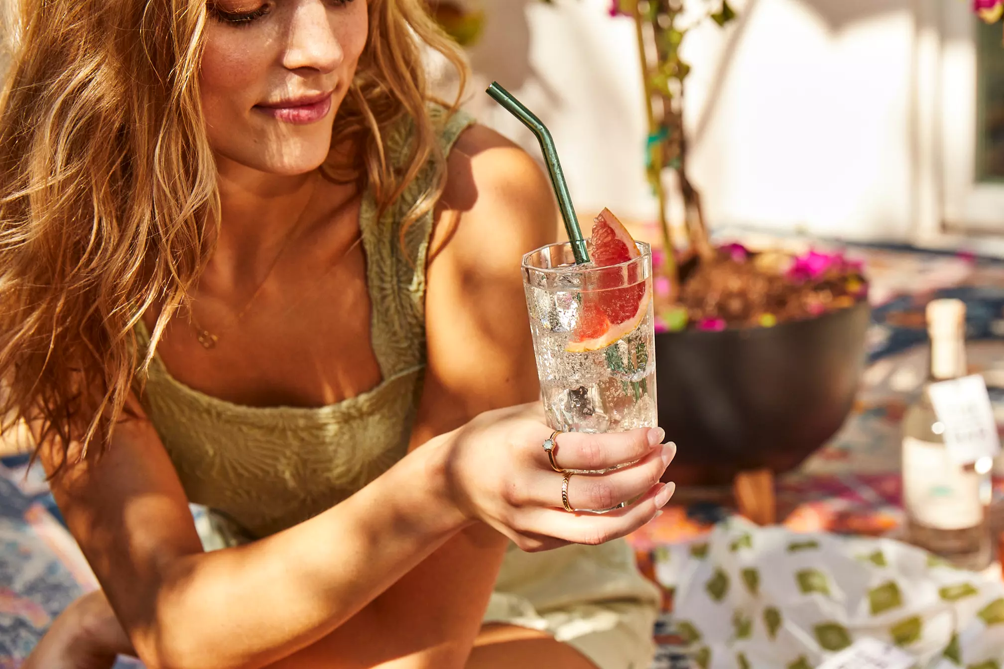 Woman holding a glass of sparkling drink with a grapefruit slice and green straw, sitting indoors on a patterned rug near a potted plant.
