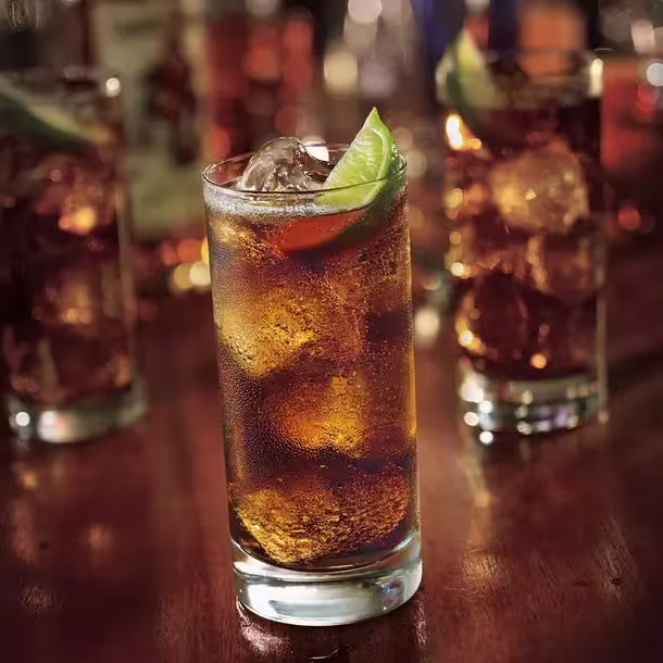 A glass of dark soda with ice and a lime wedge sits on a wooden surface, with similar drinks visible in the background.