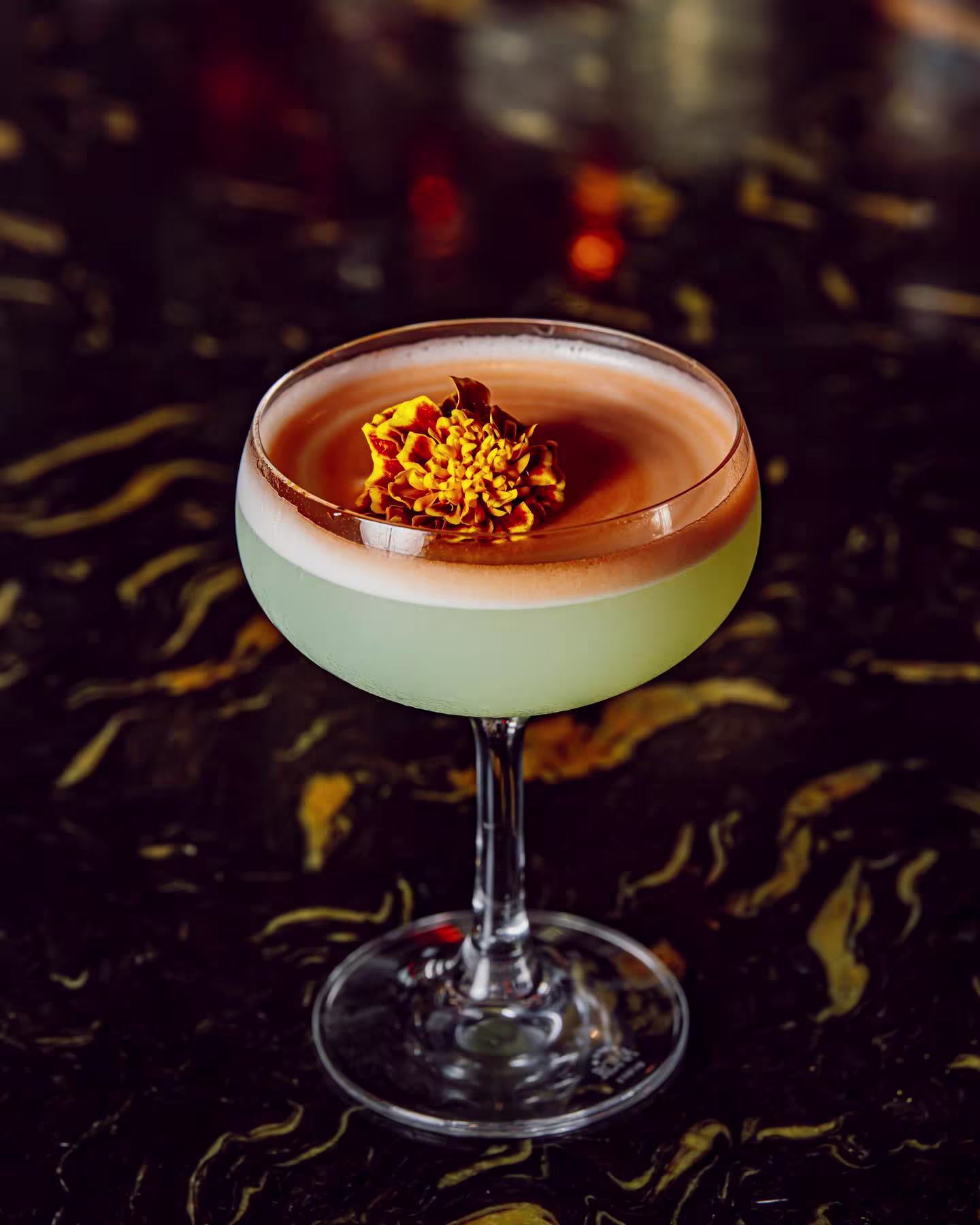 En Gangtas, un vibrante cóctel verde con espuma beige y una flor naranja flotando, destaca sobre una elegante barra de mármol oscuro.