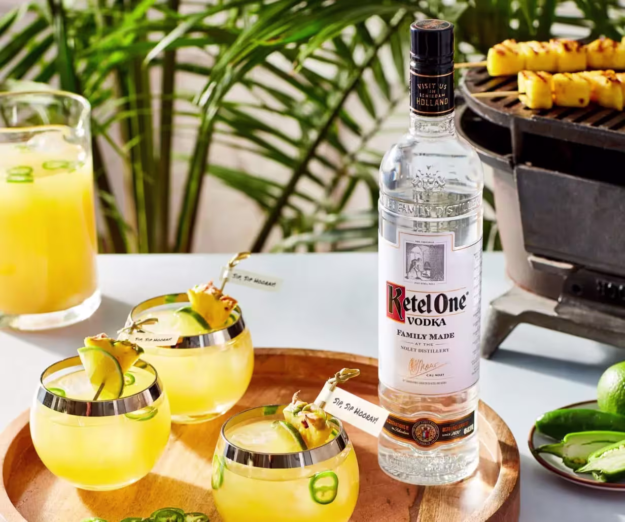 A bottle of Ketel One vodka sits on a table with three yellow cocktails garnished with pineapple and jalapeño, a pitcher of juice, limes, and a grill with pineapple skewers in the background.
