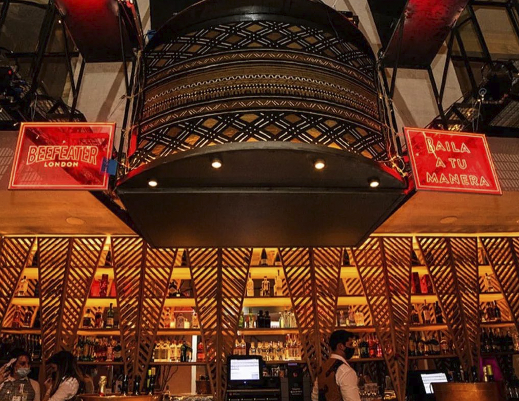 Interior de La Reserva con barra iluminada, diseño decorativo en madera y carteles rojos que dicen 'Baila a tu manera' y 'Beefeater London'