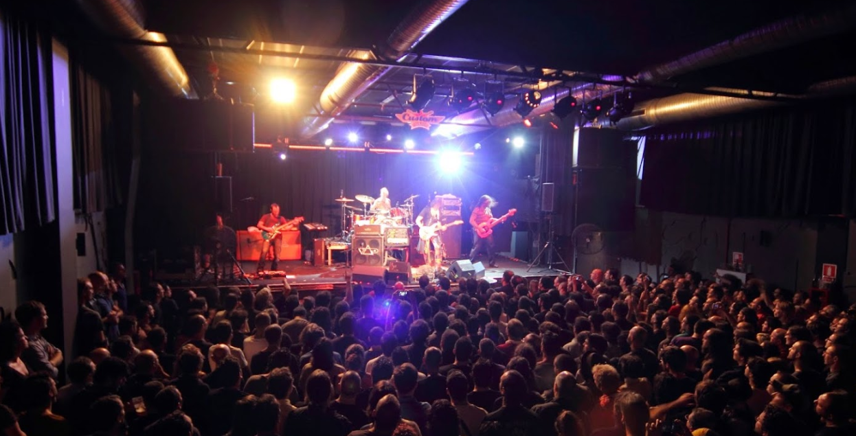La imagen muestra una actuación de música en la Sala Custom. Una banda está en el escenario con varios instrumentos, incluyendo guitarras y baterías, iluminados por luces de escenario.