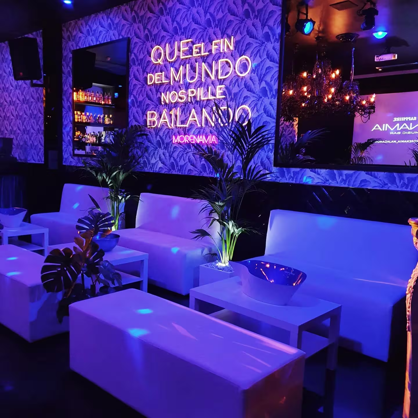 Zona lounge de Morena Mía con sofás blancos, luces azules y neón con la frase "Que el fin del mundo nos pille bailando". Ambiente chic y relajado