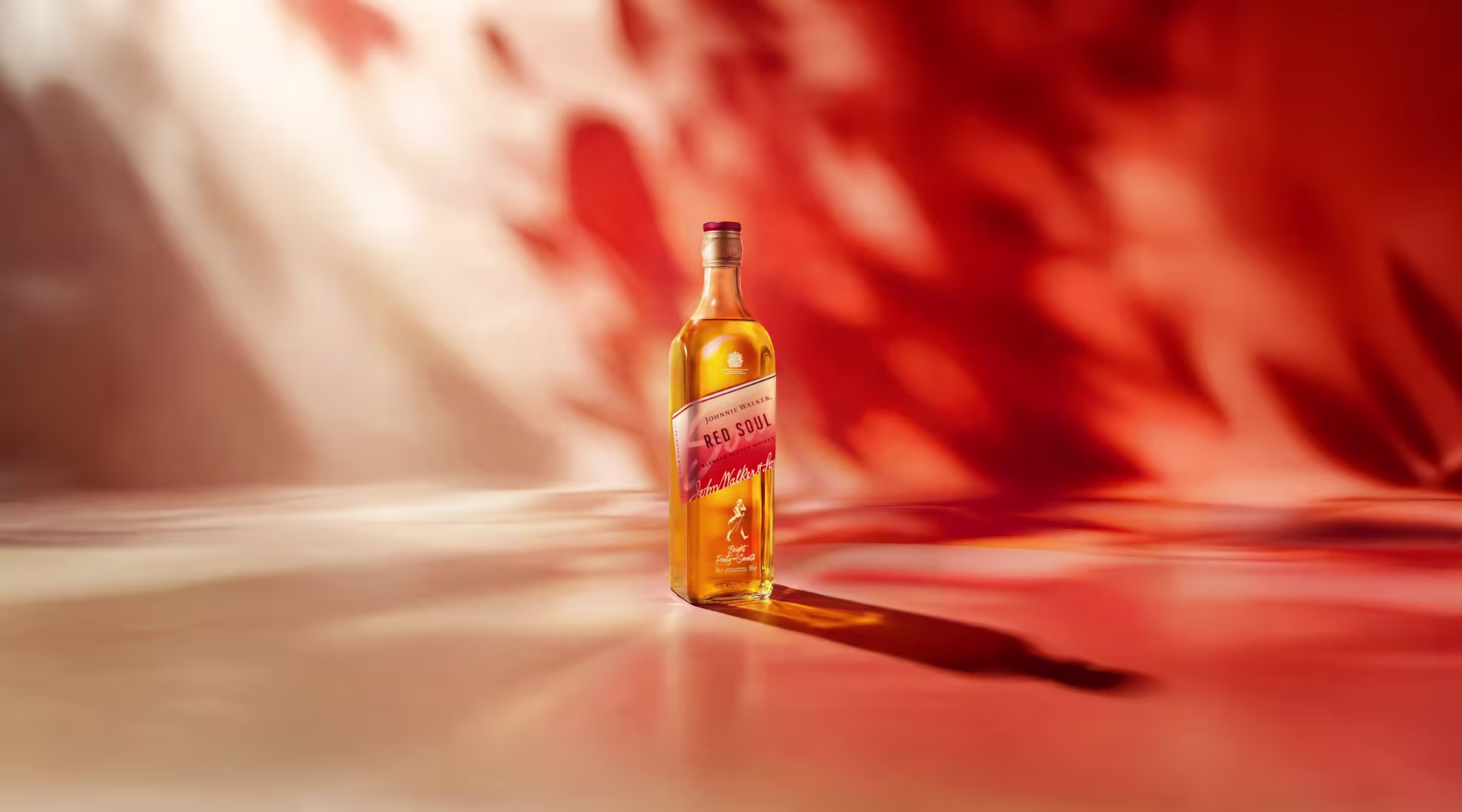 Botella de whisky Johnnie Walker Red Soul colocada en posición vertical sobre una superficie lisa, iluminada con cálidos tonos dorados, con un fondo abstracto difuminado en tonos rojos y naranjas y una larga sombra proyectada hacia la derecha.