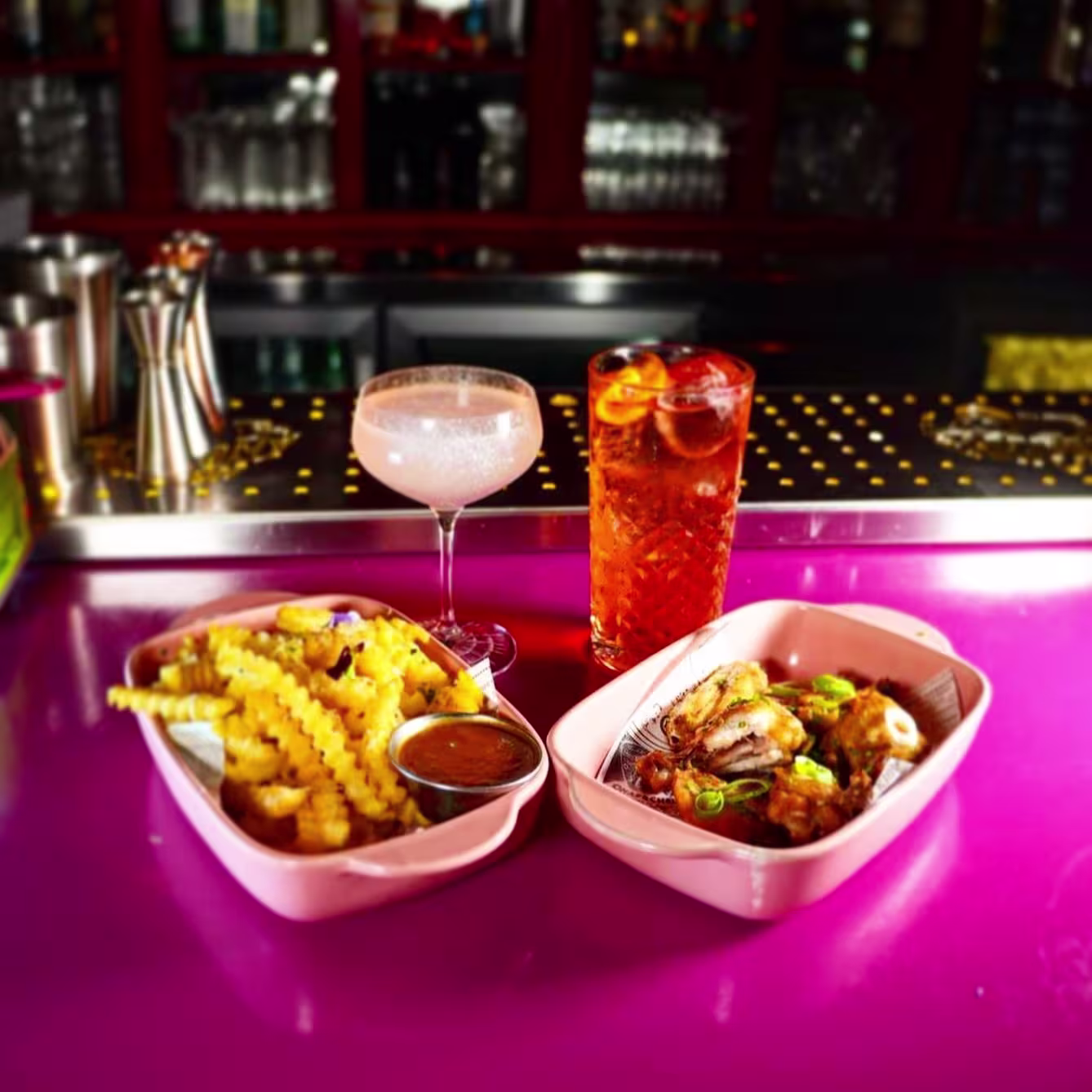 La foto muestra un entorno Love Cocktail Company, con un enfoque en la comida y las bebidas colocadas sobre una superficie vibrante de color rosa. Hay dos platos de comida que lo acompañan.