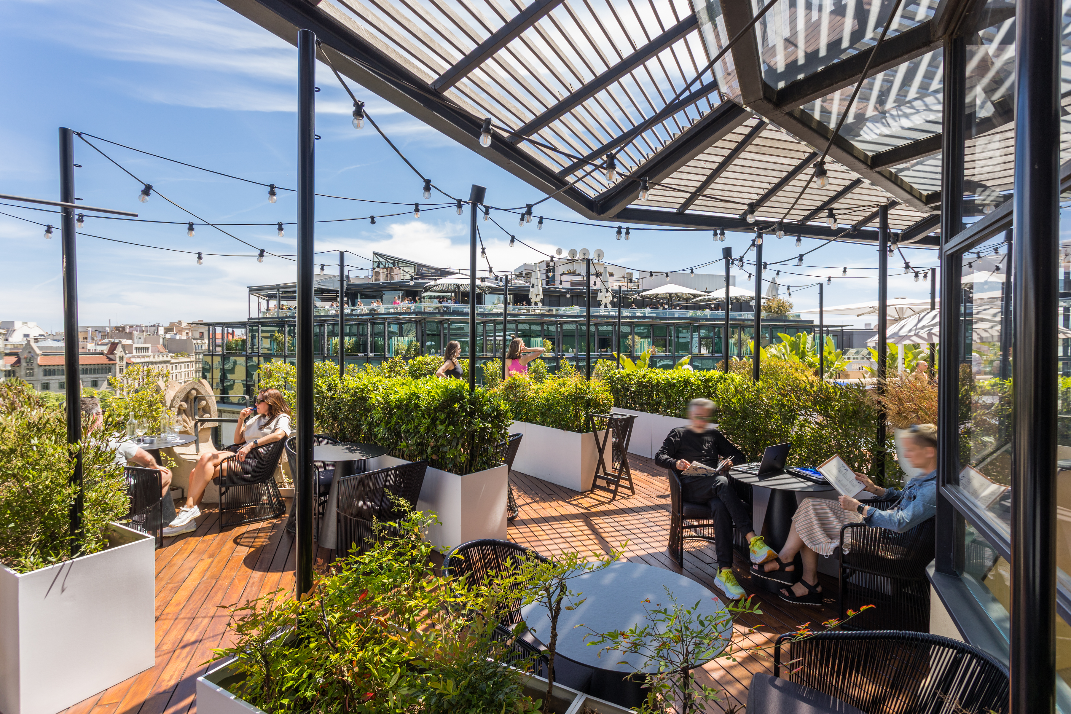 La imagen muestra una terraza en la azotea Verbena, con una estructura de vidrio y metal. Hay varias personas sentadas en diferentes mesas. La terraza está adornada con plantas en macetas y luces colgantes.