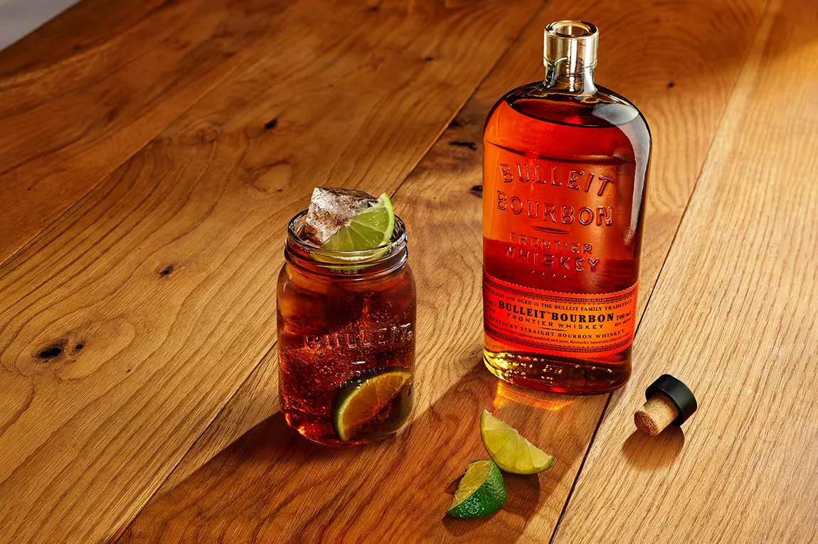 A bottle of Bulleit Bourbon whiskey, a glass with ice and lime wedges, and a cork on a wooden surface.