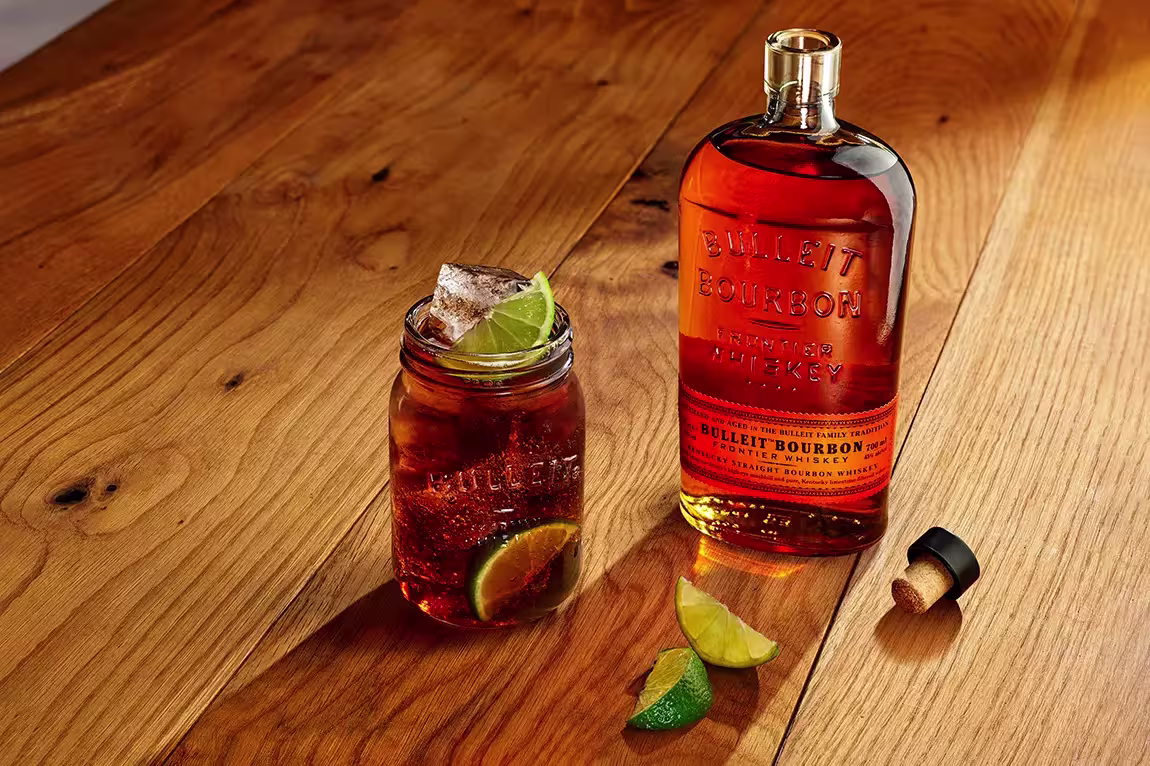 A bottle of Bulleit Bourbon whiskey, a glass with ice and lime wedges, and a cork on a wooden surface.